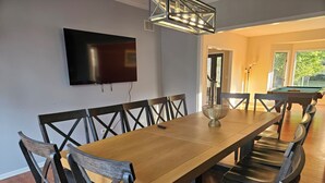 A dining area with a long wooden table, dark wooden chairs, a television mounted on the wall, and a pool table in the background. - Fun 5BR 4.5BA, Pool, Hot Tub, Game Rm, Near O’Hare (Wood Dale)
