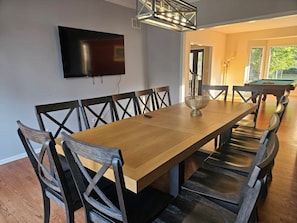 A dining area with a long wooden table, dark wooden chairs, a television mounted on the wall, and a pool table in the background. - Fun 5BR 4.5BA, Pool, Hot Tub, Game Rm, Near O’Hare (Wood Dale)