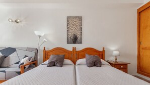 Una cama doble con cabecera de madera, una mesita de noche con lámpara, una obra de arte en la pared y un sofá con estampado geométrico.