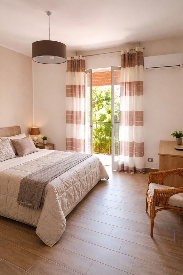 A bedroom with a bed, a bedside table with a lamp, a wicker chair, and a view through curtains to a balcony.