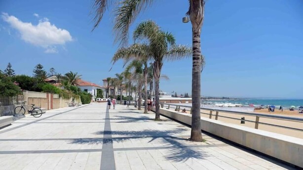 Eine gepflasterte Promenade mit Palmen, ein Strand im Hintergrund und ein klarer, blauer Himmel.
