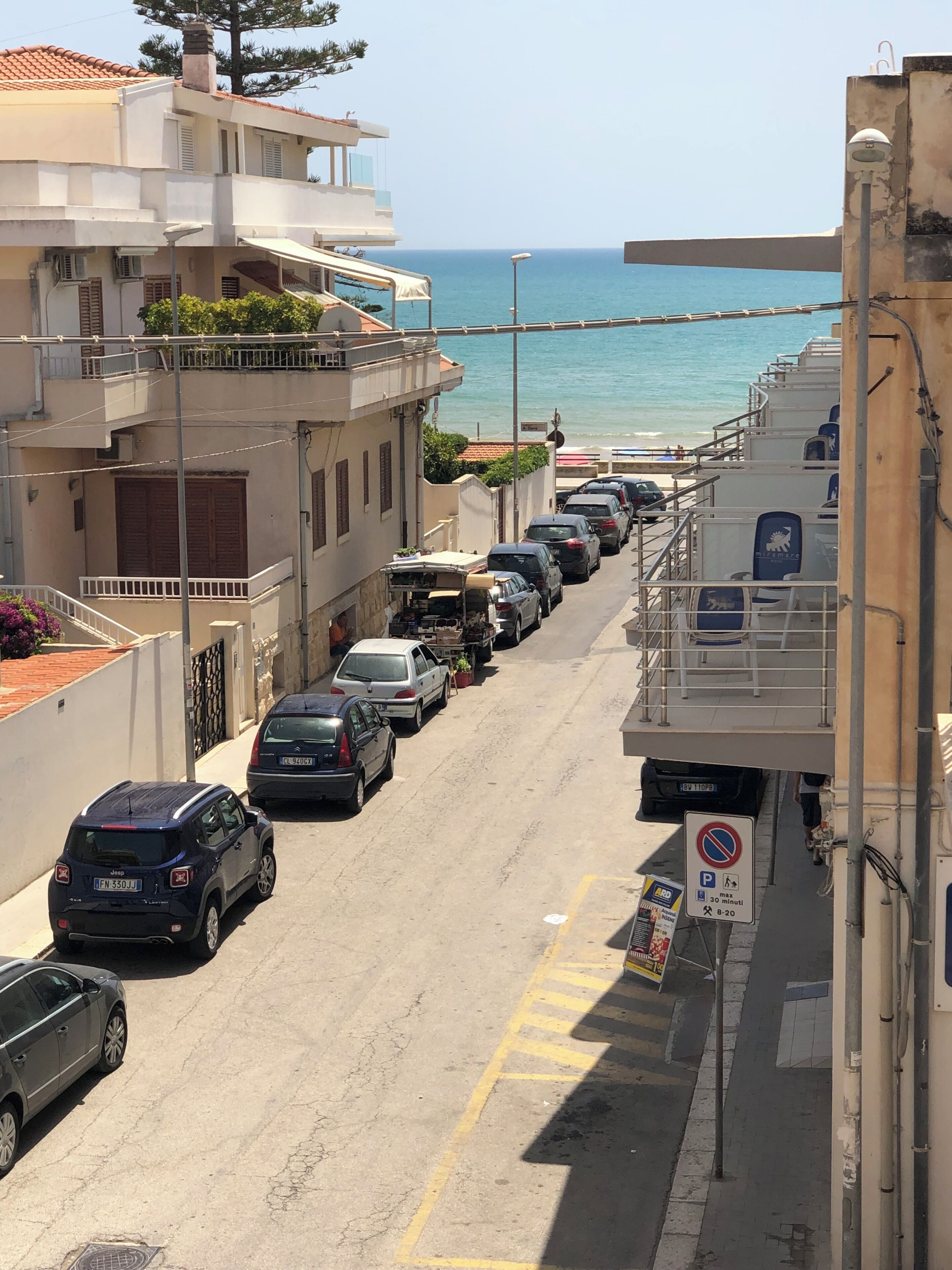 A coastal street lined with parked cars and buildings, including a visible balcony and a no parking sign.