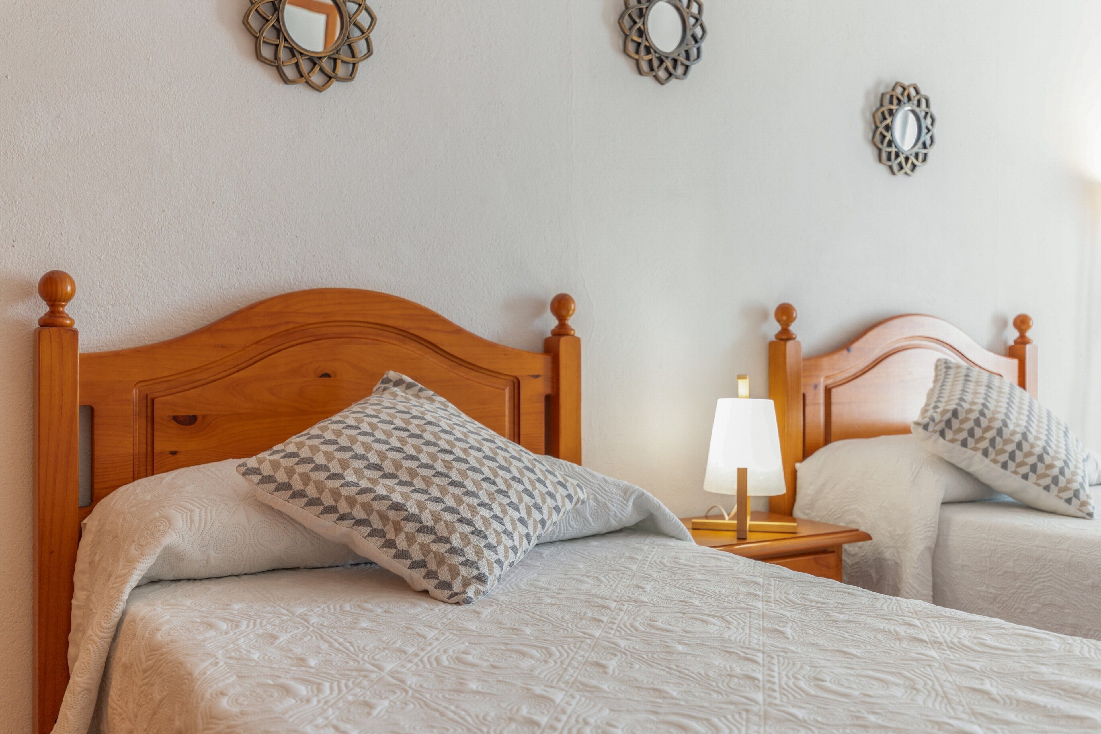 Una cama doble con cabecera de madera, un cojín estampado, una mesita de noche con lámpara y espejos decorativos en la pared.