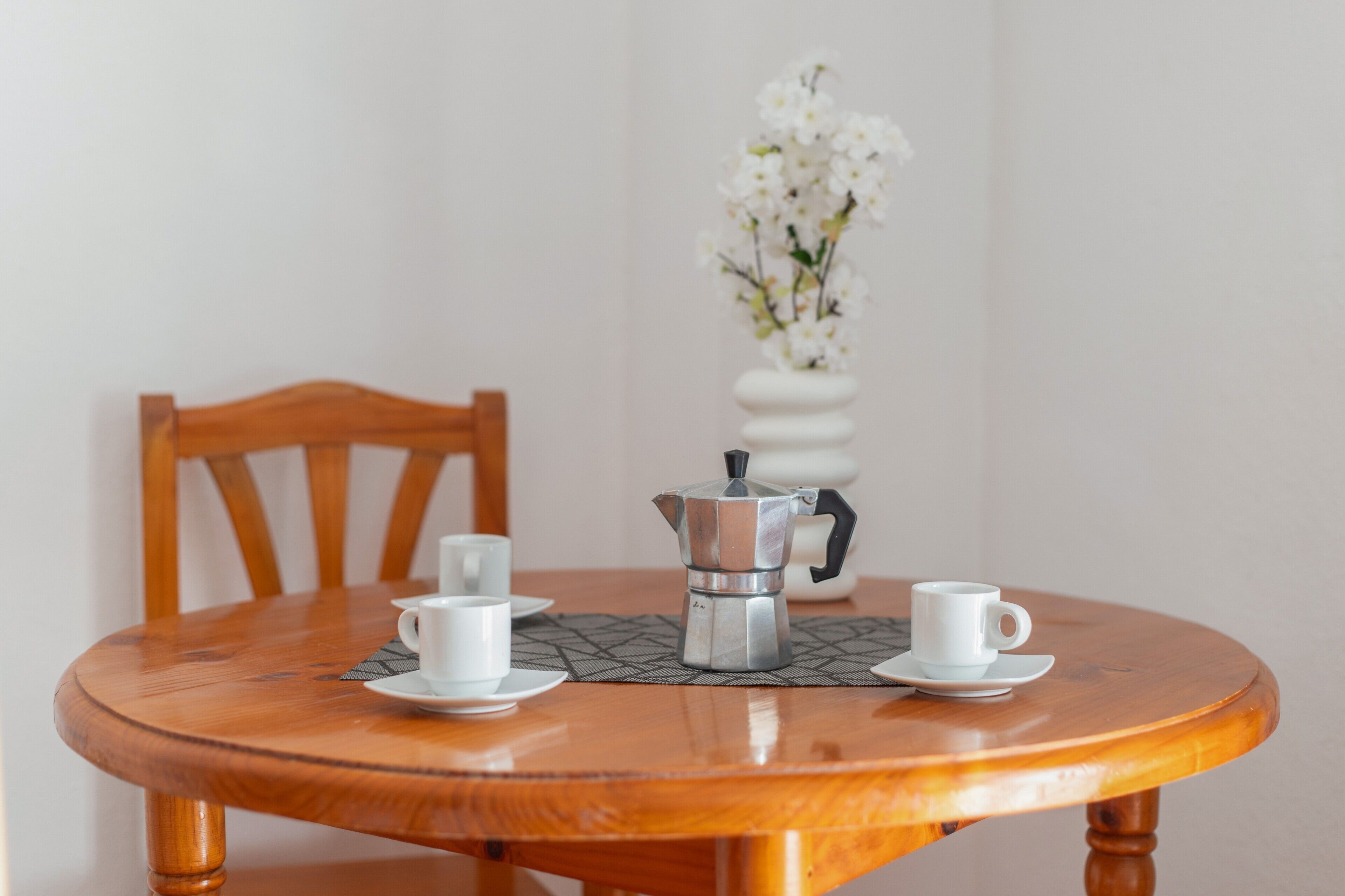 Une table en bois avec une cafetière et des tasses, un vase rempli de fleurs blanches et une chaise.
