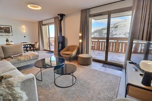 A modern living room with a sofa, two glass tables, a chair, a fireplace, and a balcony with a view of a snowy mountain.