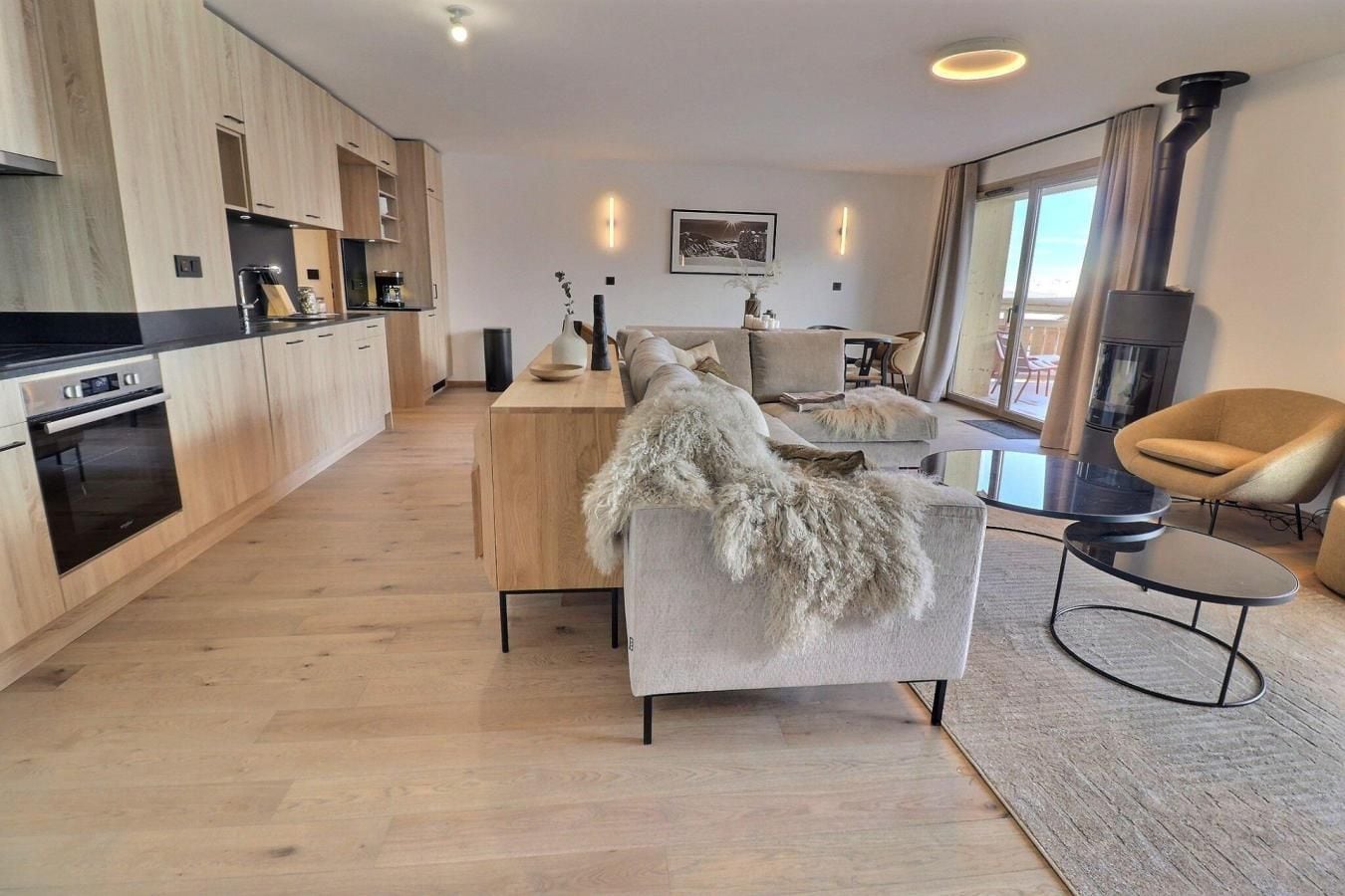 A modern living space with a kitchen, a sofa with a fur throw, a round coffee table, and a fireplace.