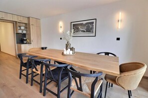A modern dining area with a wooden table and six chairs, a kitchen area with wooden cabinets, and a framed picture on the wall. - 30m from ski slopes, 1st floor, view mountain, terrace, fireplace or stove, 134m², Méribel (Les Allues)