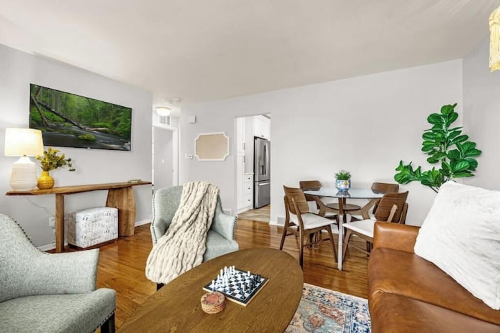 A modern living room with a sofa, a coffee table with chess pieces, a dining area with chairs, a kitchen area, and a large window with a view of greenery.