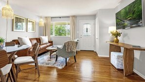 A modern living room with a wooden floor, a leather sofa, a dining table with chairs, a coffee table, and a TV on the wall. - New Adventure Cottage w/ Mountain Views Near Garden of the God's and much more! (Colorado Springs)