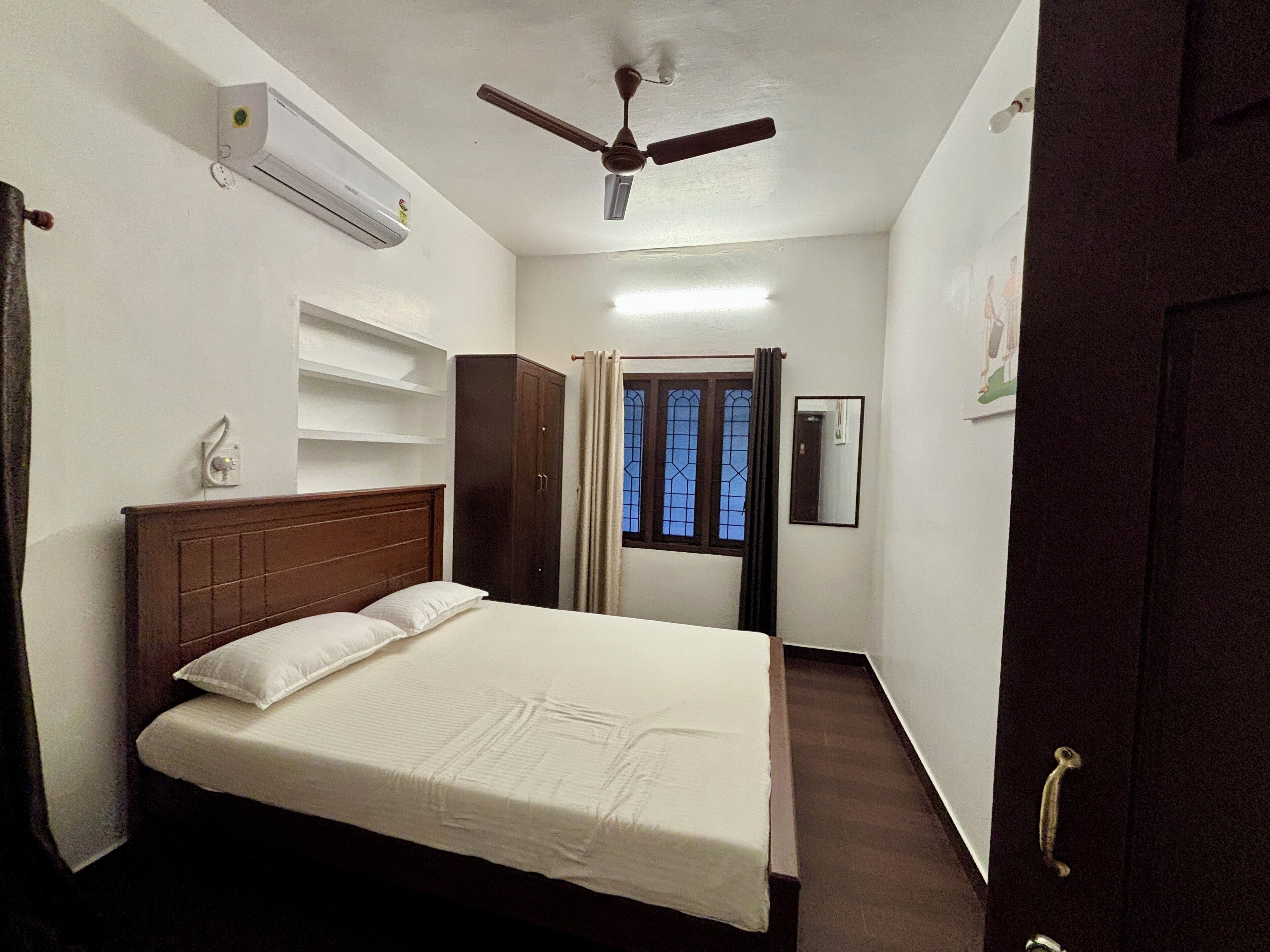 A bedroom with a bed, a wardrobe, a ceiling fan, an air conditioning unit, a window with curtains, and a door.