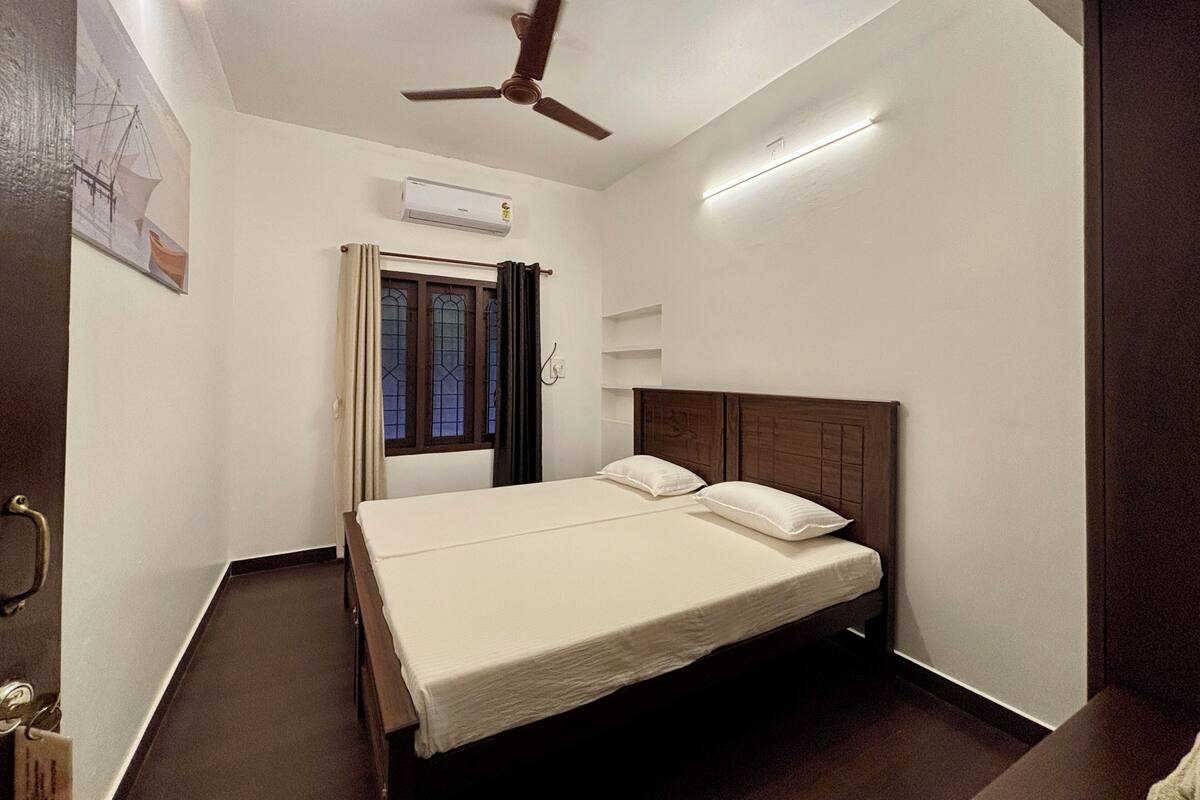 Un dormitorio con cama, ventilador de techo, ventana con cortinas y equipo de aire acondicionado.