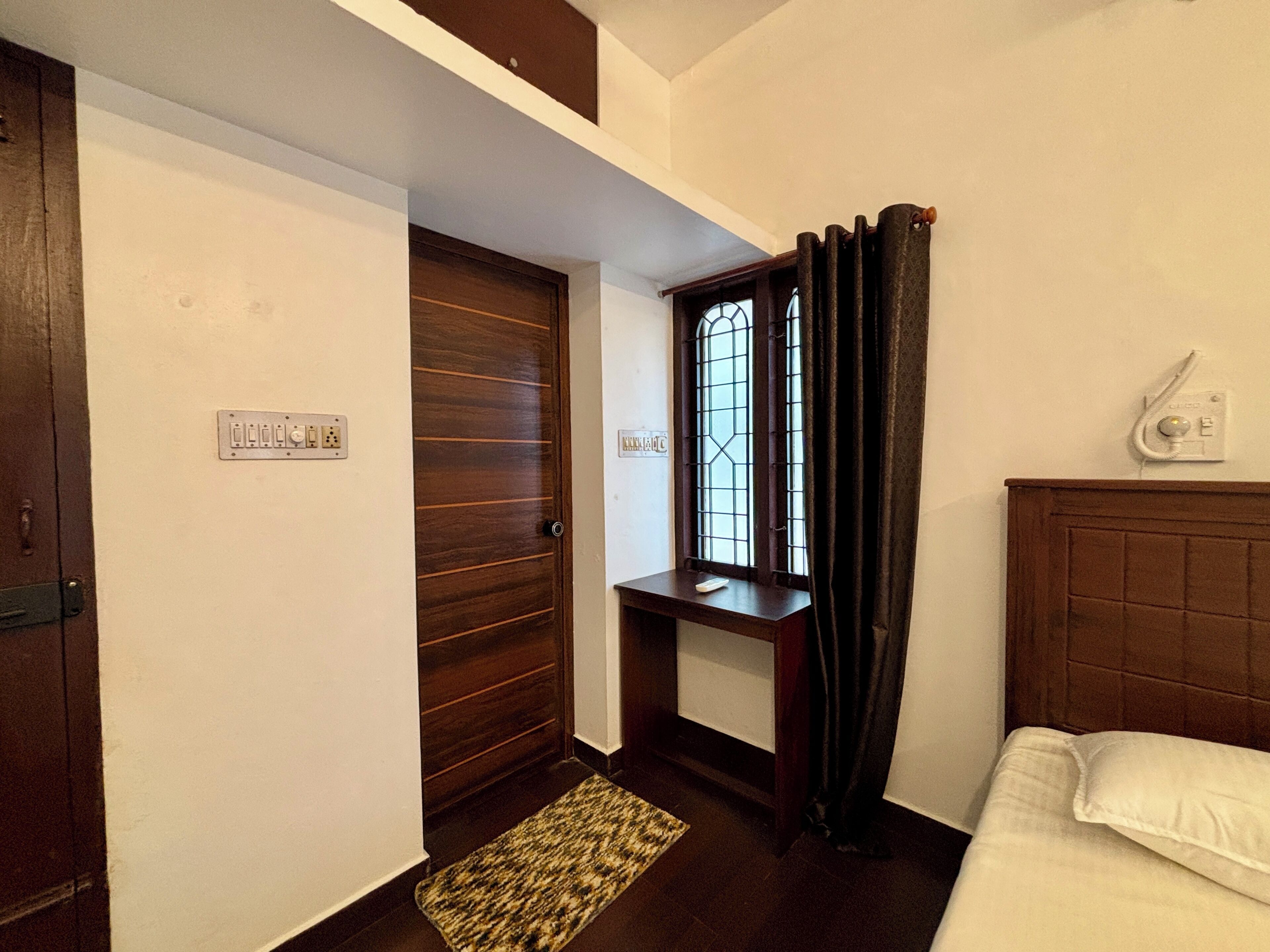 Una habitación de hotel con puerta de madera, una mesita, una ventana con cortinas, una mesita de noche y una cama con almohada blanca.