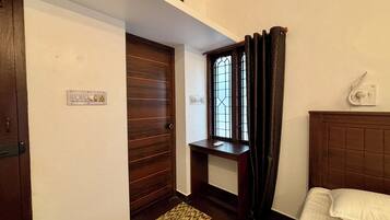 Una habitación de hotel con puerta de madera, una mesita, una ventana con cortinas, una mesita de noche y una cama con almohada blanca.