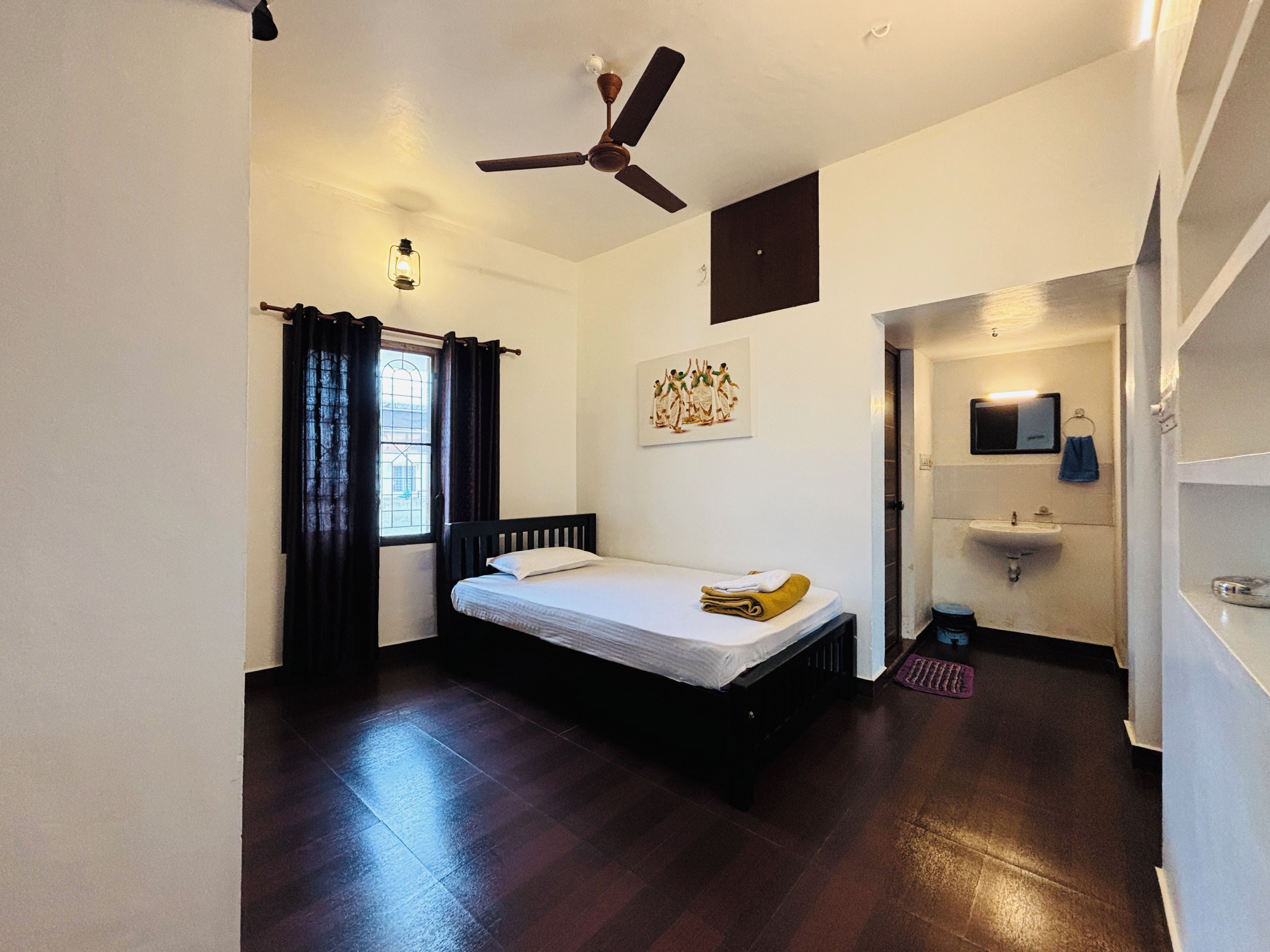 Une chambre à coucher avec un lit, un ventilateur de plafond, une fenêtre avec des rideaux, un tableau au mur et une salle de bain avec un lavabo et un miroir.