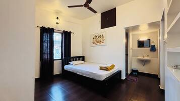 Une chambre à coucher avec un lit, un ventilateur de plafond, une fenêtre avec des rideaux, un tableau au mur et une salle de bain avec un lavabo et un miroir.