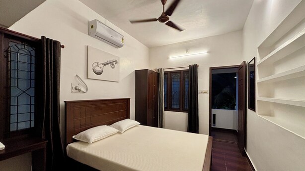 A bedroom with a bed, an air conditioning unit, a ceiling fan, a window with curtains, and a door leading to a balcony. - Fort Grande Stay  (Kochi)