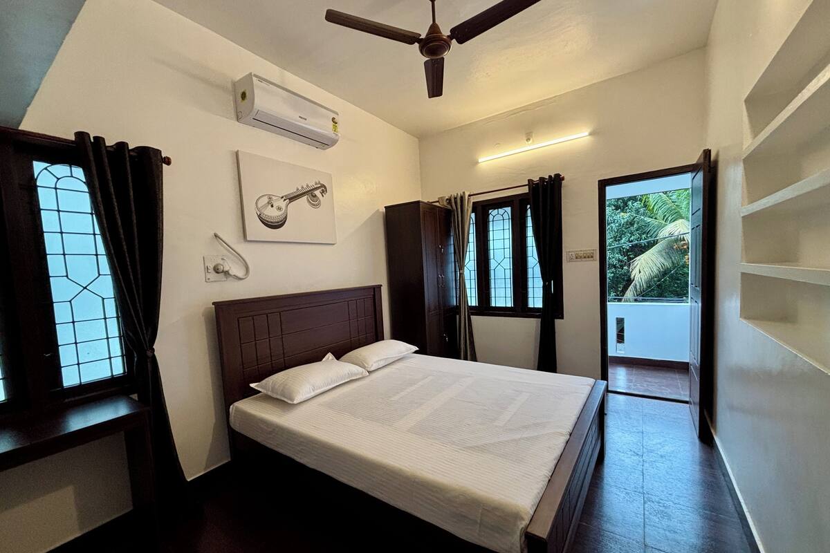 Un dormitorio con cama, aire acondicionado, ventilador de techo, ventana con cortinas, una lámpara de pared y una puerta que da a un balcón.