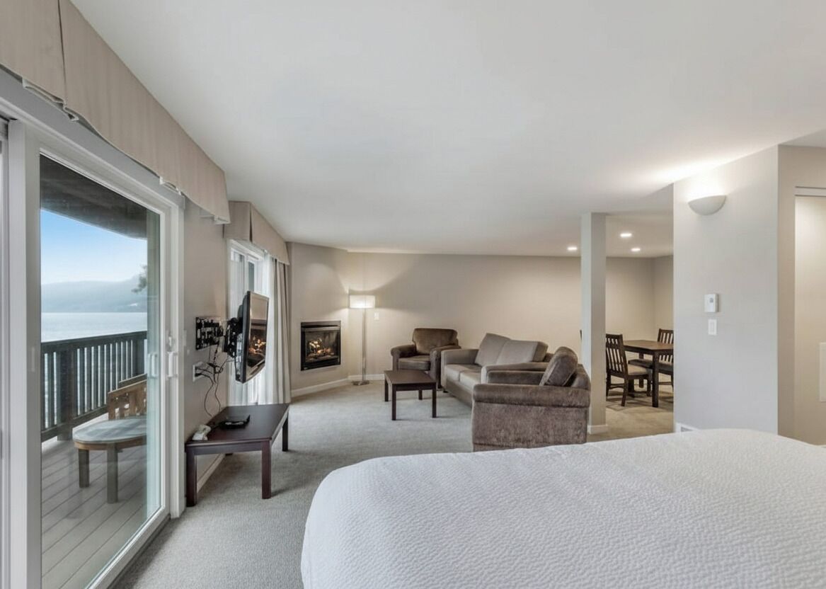 A hotel room with a large bed, a sofa, a small table, and a TV. There is a balcony with a view of the sea.