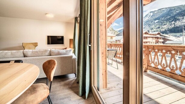 A modern living room with a sofa, a dining table, and chairs. A large window offers a view of a balcony with a wooden railing and a mountainous landscape. - 100m from resort center, 2nd floor, view village, terrace, tv, 84m², Morzine (Morzine)
