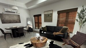 Een moderne woonkamer met een ronde houten salontafel, een zwarte leren bank, een eethoek met een witte tafel en stoelen, en een groot abstract schilderij aan de muur.