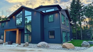 A modern two-story house with large windows and a wooden front door, surrounded by rocks and trees. - Newly Built Luxury Chalet Indoor Pool Ski Lift (The Blue Mountains)