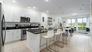 Een moderne keuken met witte kasten, een centraal eiland met barstoelen, een zithoek met tafel en stoelen, en uitzicht op groen door grote ramen.