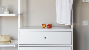 A white wardrobe with a hanging rod, a drawer, and two apples on top.
