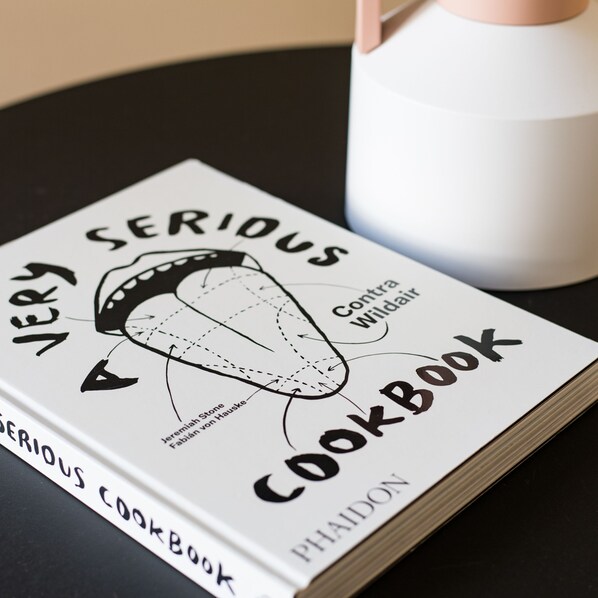 A cookbook titled 'Very Serious Cookbook' by Phaidon, placed on a table next to a white pitcher. - Bright Covent Garden Studio - Netflix Nespresso (London)