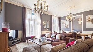 Een ruime woonkamer met een grote bruine leren bank, een houten salontafel en een televisie op een meubel. Twee kroonluchters, een grote spiegel en kunstwerken sieren de muren.