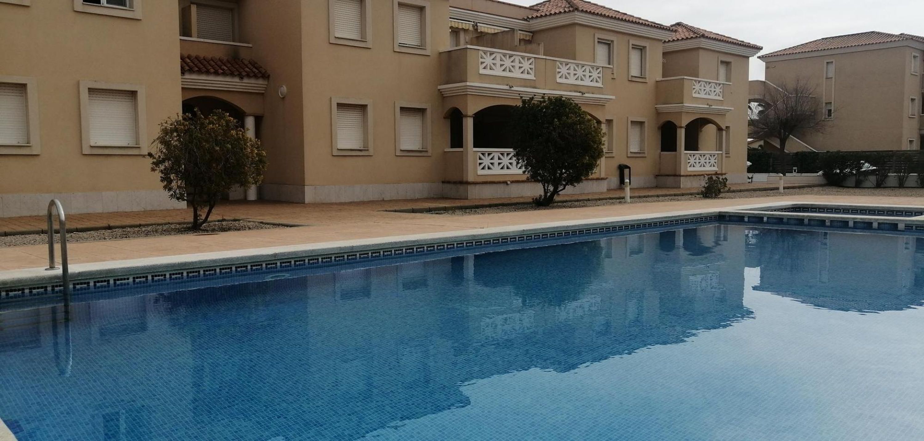 Una piscina amb una superfície d'aigua blava i transparent, envoltada per un edifici beix amb balcons i finestres.