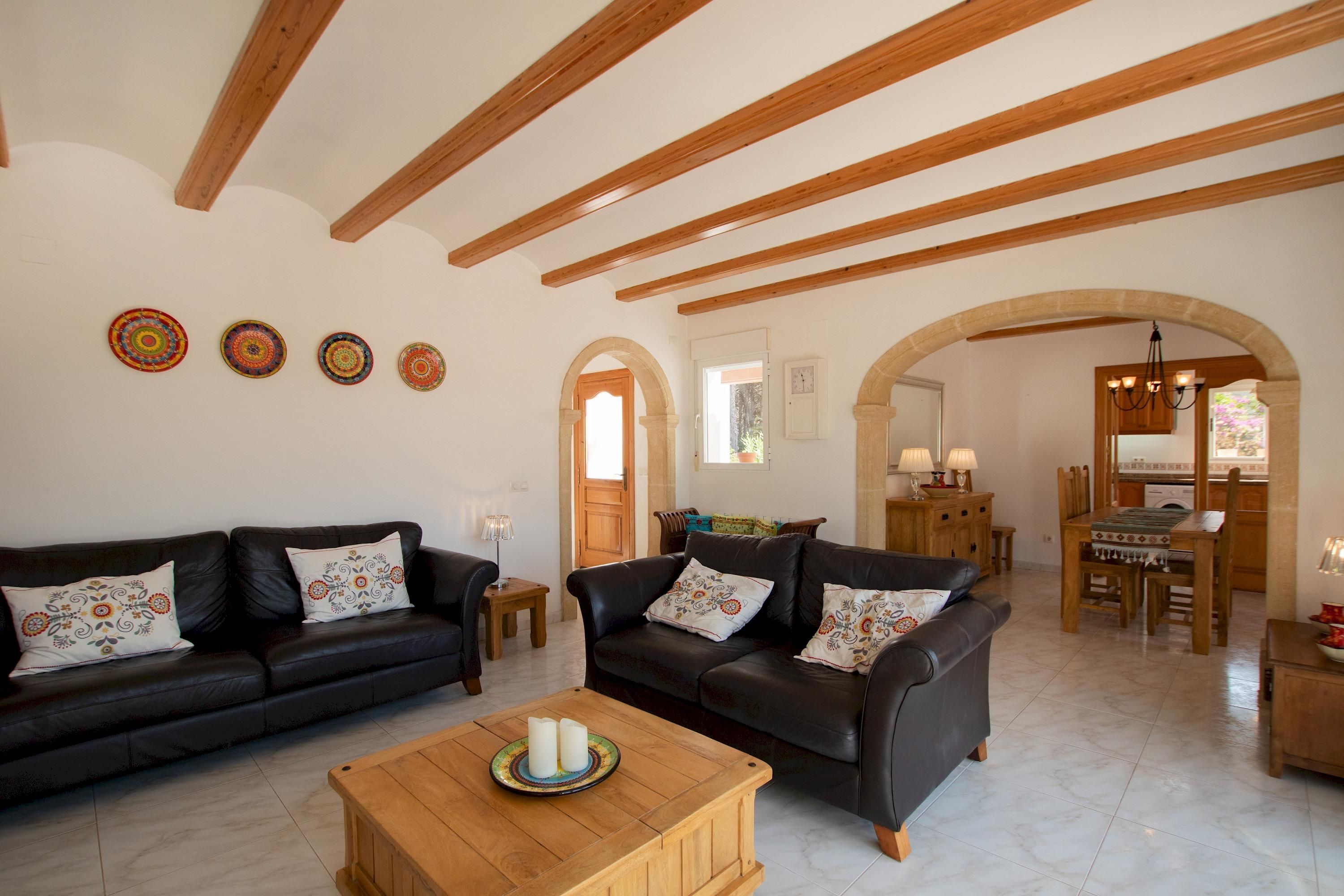 A spacious living area with two black leather sofas, a wooden coffee table, and a dining table with chairs. There are wooden beams on the ceiling and a chandelier.