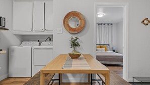 A compact kitchen with white appliances, a wooden dining table, and a view into a bedroom with a bed and curtains. - Flint A (Waco)
