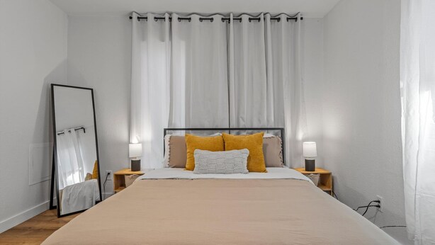 A bedroom with a large bed, two bedside lamps, a mirror, and a window with curtains. - Flint A (Waco)