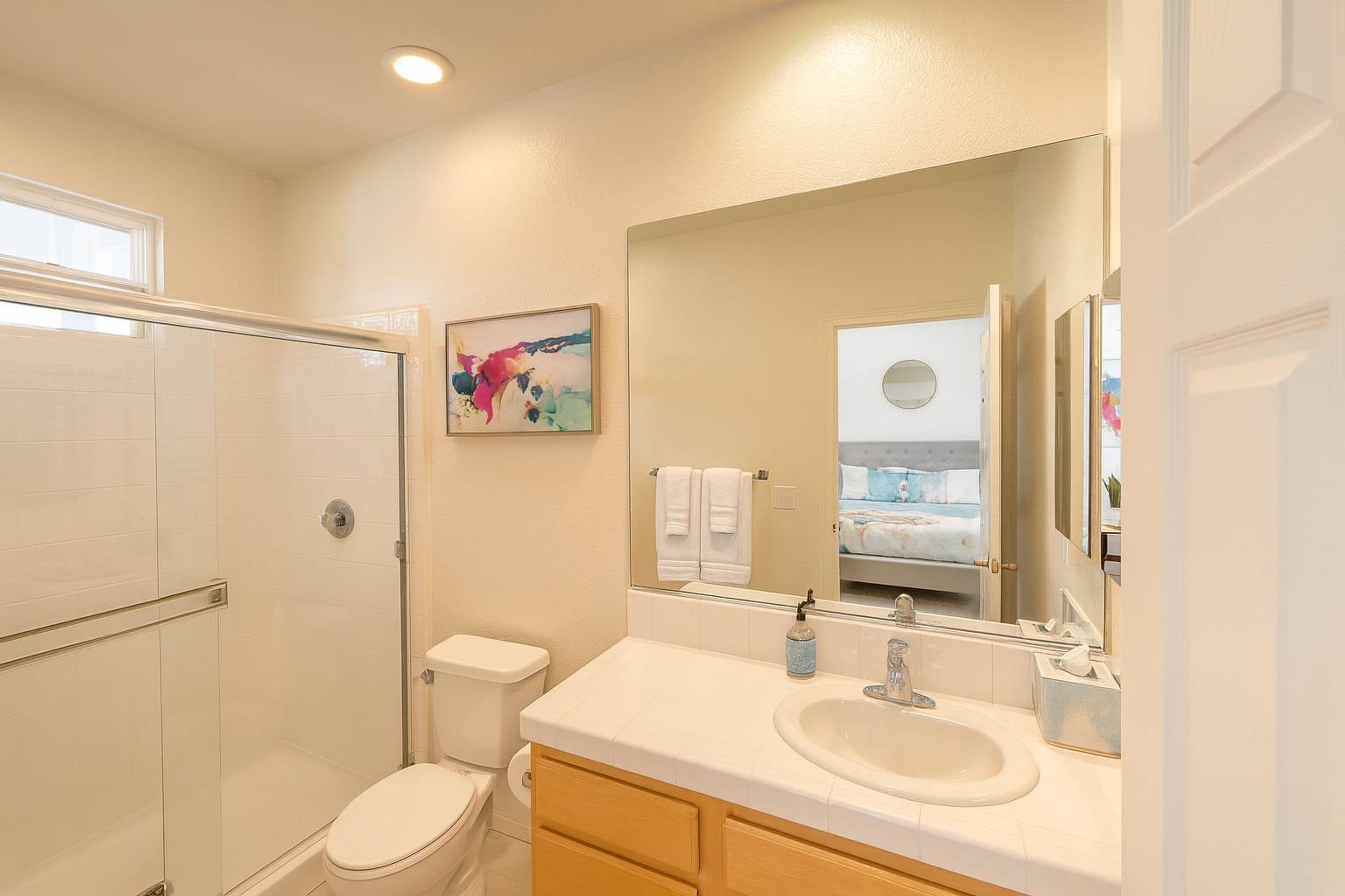 A bathroom with a glass shower enclosure, a toilet, a sink with a mirror, and a colorful artwork on the wall.