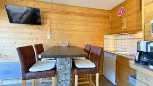 Interior de una cabaña de madera con una mesa de comedor, cuatro sillas, un televisor montado en la pared, una zona de cocina con cafetera, y un letrero que indica un precio de 15.