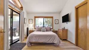 Un dormitorio con cama, televisor montado en la pared, ventana con vista a vehículos estacionados y puerta corrediza de vidrio que da a un espacio exterior.