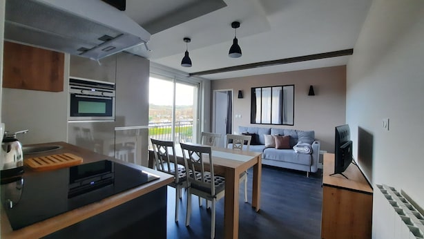 A modern kitchen with a central island, a dining area with a table and chairs, a sofa, and a television. - The magnolia (Hautot-sur-Mer)