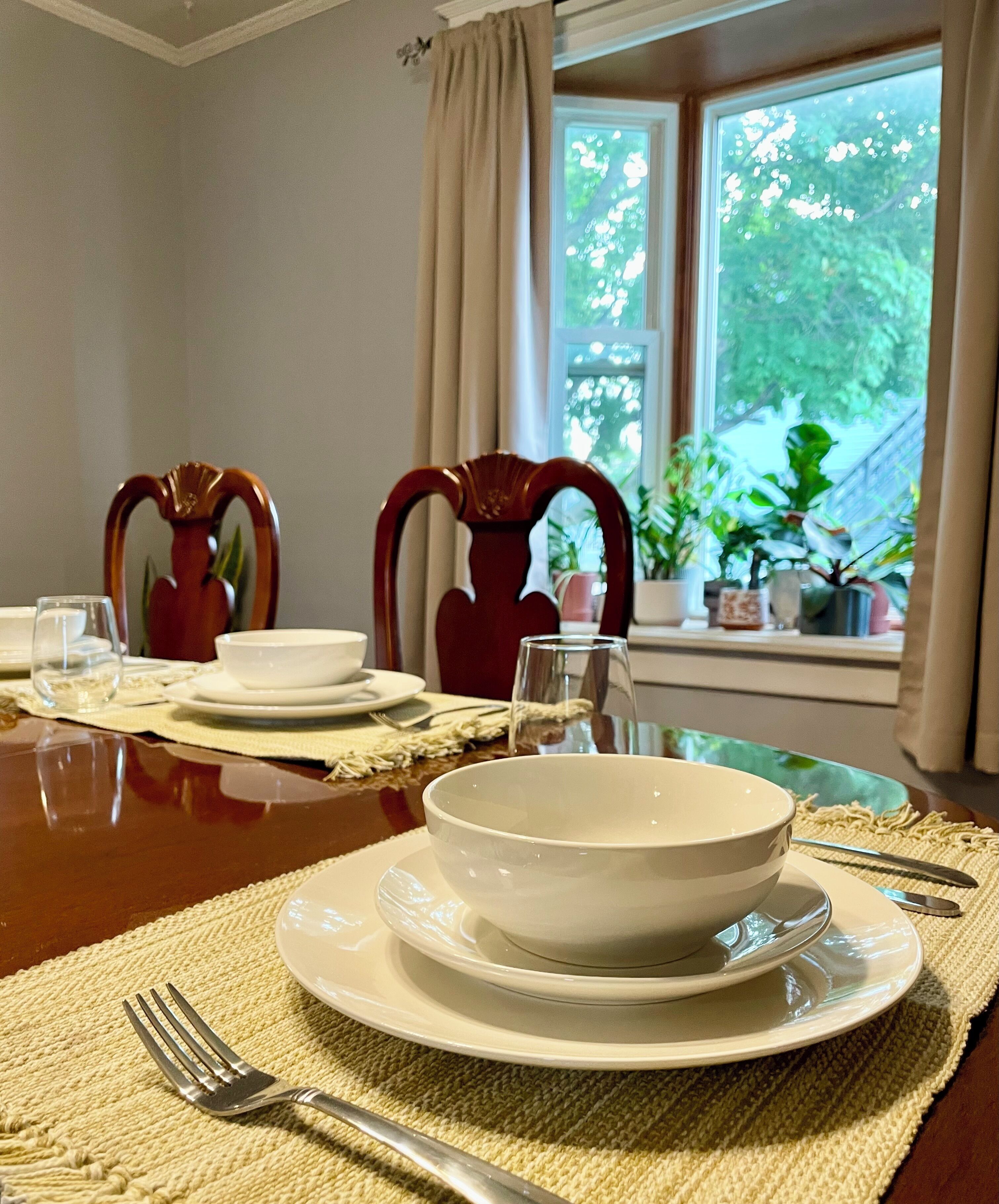 A dining table set with white plates, bowls, and cutlery, with a view of greenery outside through a window.