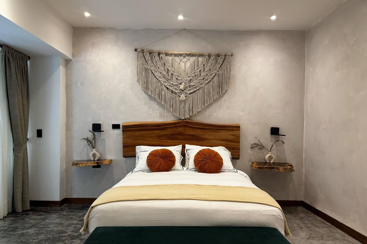 Une chambre moderne avec une tête de lit en bois, une décoration murale en macramé, des tables de chevet ornées de vases et un grand lit avec deux oreillers.