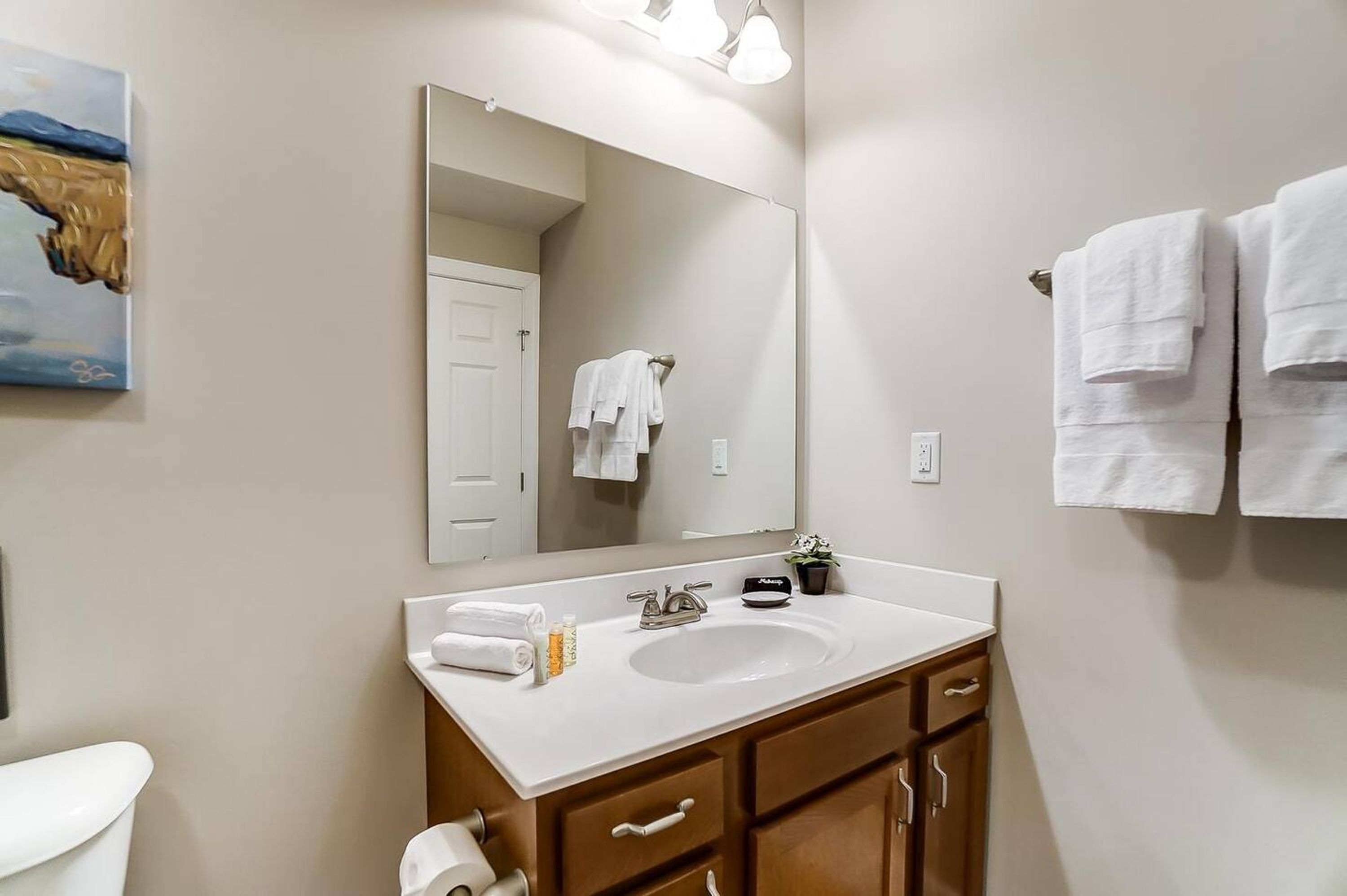 A bathroom with a white sink, a mirror, and a toilet. There are towels hanging on the wall and a painting above the sink.