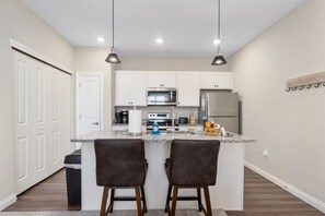A modern kitchen with white cabinets, a granite countertop, two bar stools, a stainless steel refrigerator, and a microwave. - Powell Retreat Attached Garage Walk-in Shower (Powell)