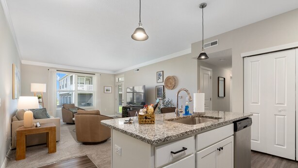 A modern kitchen with granite countertops, white cabinetry, and a dishwasher. There is a dining area with a sofa and a lamp. - Powell Retreat Attached Garage Walk-in Shower (Powell)