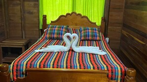 A wooden bed with a colorful striped bedspread and pillows, a green curtain, and a wooden headboard. - Green Jay Cabin 2 (Cristo Rey)
