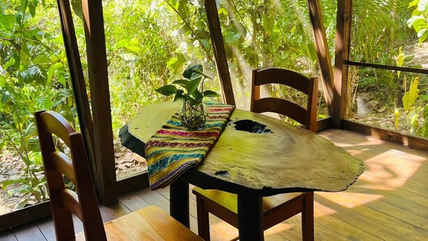 An outdoor wooden table with chairs, surrounded by a wooden pergola with a view of dense green foliage. - Green Jay Cabin 2 (Cristo Rey)