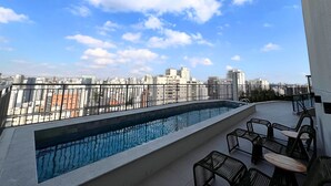 Une piscine sur le toit offrant une vue sur la ville, entourée d’une rambarde en métal et agrémentée de mobilier d’extérieur.