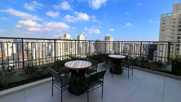 Une terrasse sur le toit, dotée de tables et de chaises rondes, entourée d’une rambarde en métal, et offrant une vue sur le paysage urbain avec ses nombreux immeubles de grande hauteur.