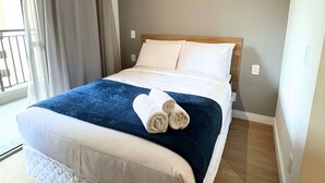 Un lit bien fait, avec une couverture bleue et des serviettes enroulées, une tête de lit en bois et une fenêtre avec des rideaux.