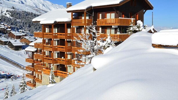 Una cabaña de madera de varios pisos con balcones, rodeada de montañas y árboles cubiertos de nieve.