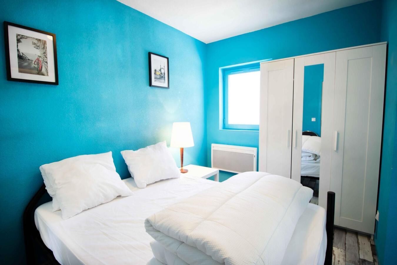 A bedroom with a bed, white bedding, a nightstand with a lamp, a window, and a wardrobe.