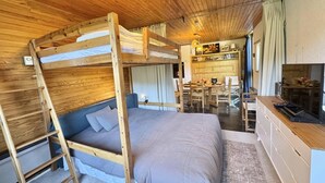Una cama litera con escalera, un sofá, un escritorio y un televisor en una habitación con techos de madera y una zona de cocina al fondo.